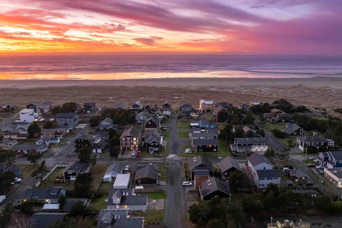 This image captures a tranquil scene with the sun setting over the ocean, houses lining a quiet seaside street, and the sandy beach stretching out toward the horizon. The neighborhood's proximity to the beach is clearly visible, making it easy to imagine stepping outside and heading straight to the shore.