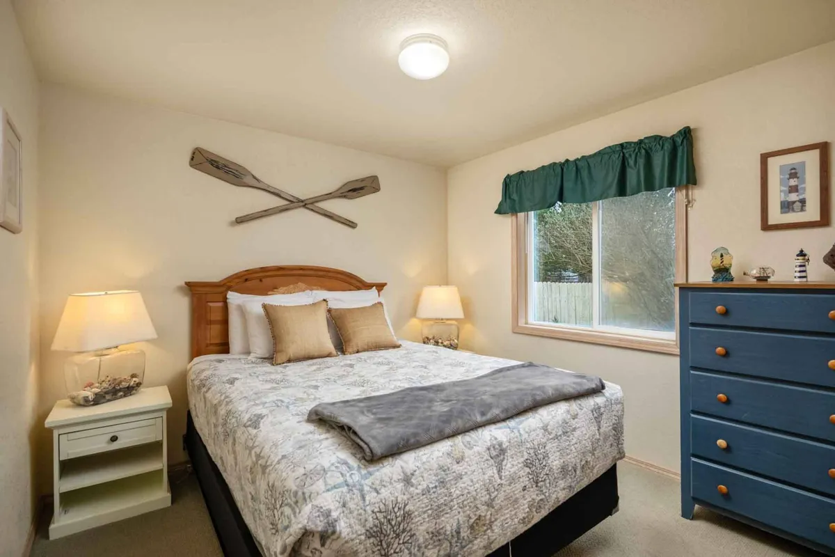 A comfortable bedroom with a bed dressed in decorative pillows, a blue dresser, and a window with a green valance that lets in soft natural light. The overall mood is quiet and serene, ideal for restful sleep or quiet mornings.