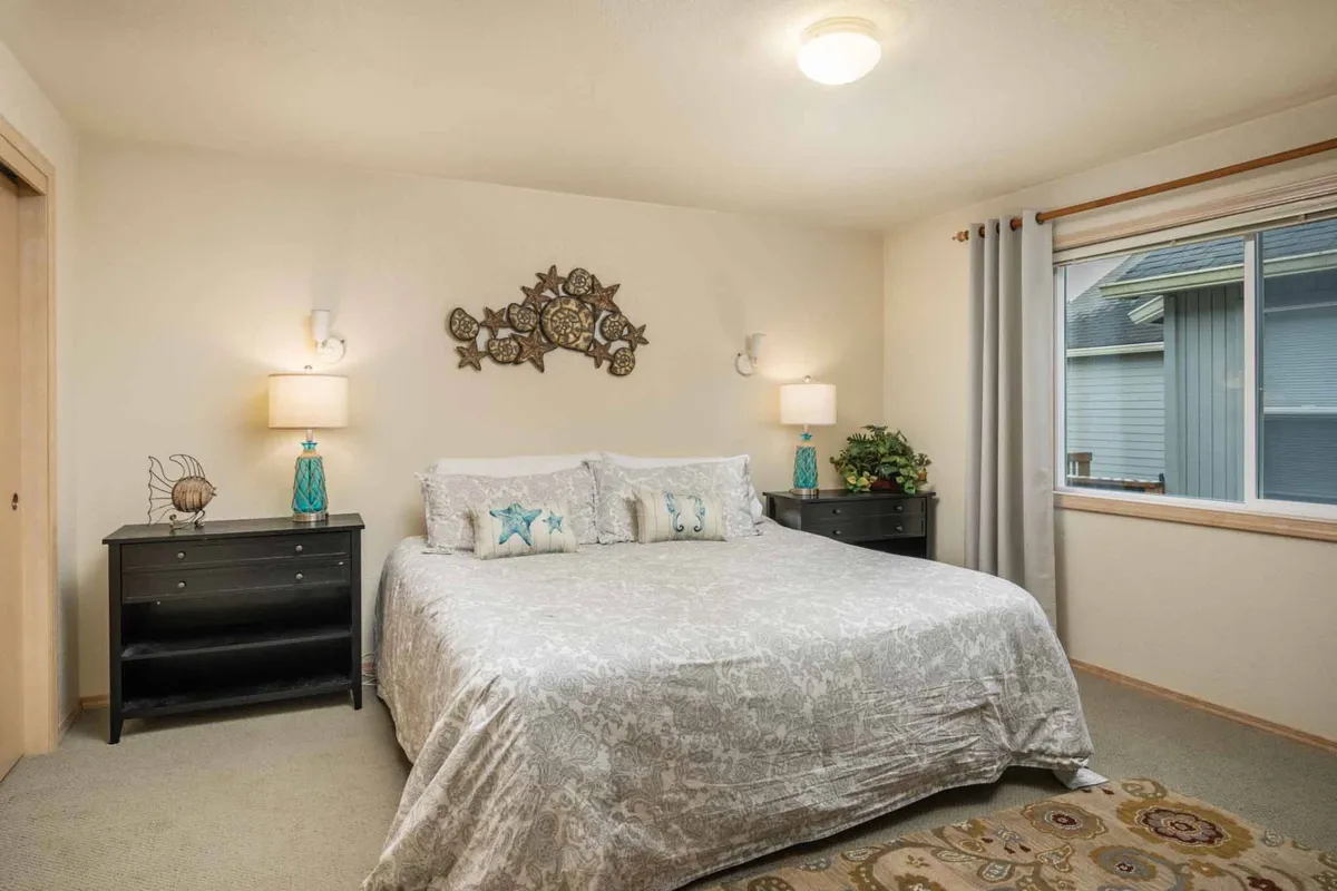 A sunlit bedroom featuring a comfortable bed adorned with decorative pillows, complemented by tasteful wall art and matching nightstands with lamps on either side. Natural light streams in, creating a warm, restful atmosphere ideal for unwinding.