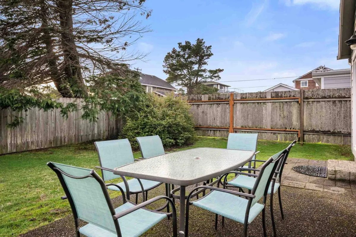 This image showcases a cheerful fenced backyard with a large outdoor dining table and chairs situated on a lush green lawn. Tall trees provide shade and a sense of privacy. The setting looks perfect for family gatherings and outdoor meals, with plenty of open space for kids to play and adults to relax.