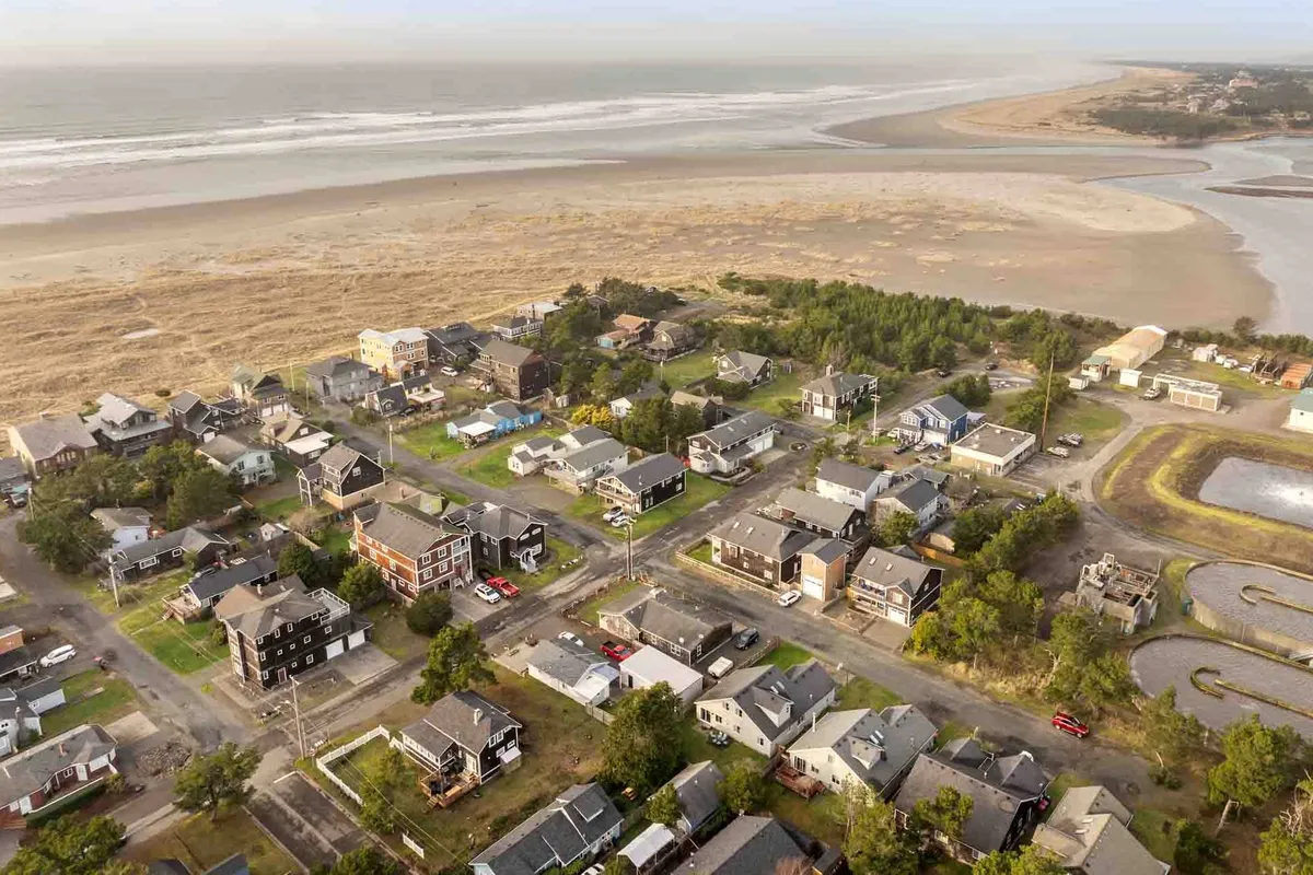 This image presents a serene coastal view with the beach in the foreground, residential homes lining the shore, and a quiet road meandering through the landscape. The atmosphere is peaceful, suggesting opportunities for beach walks and connection with nature right outside the house.