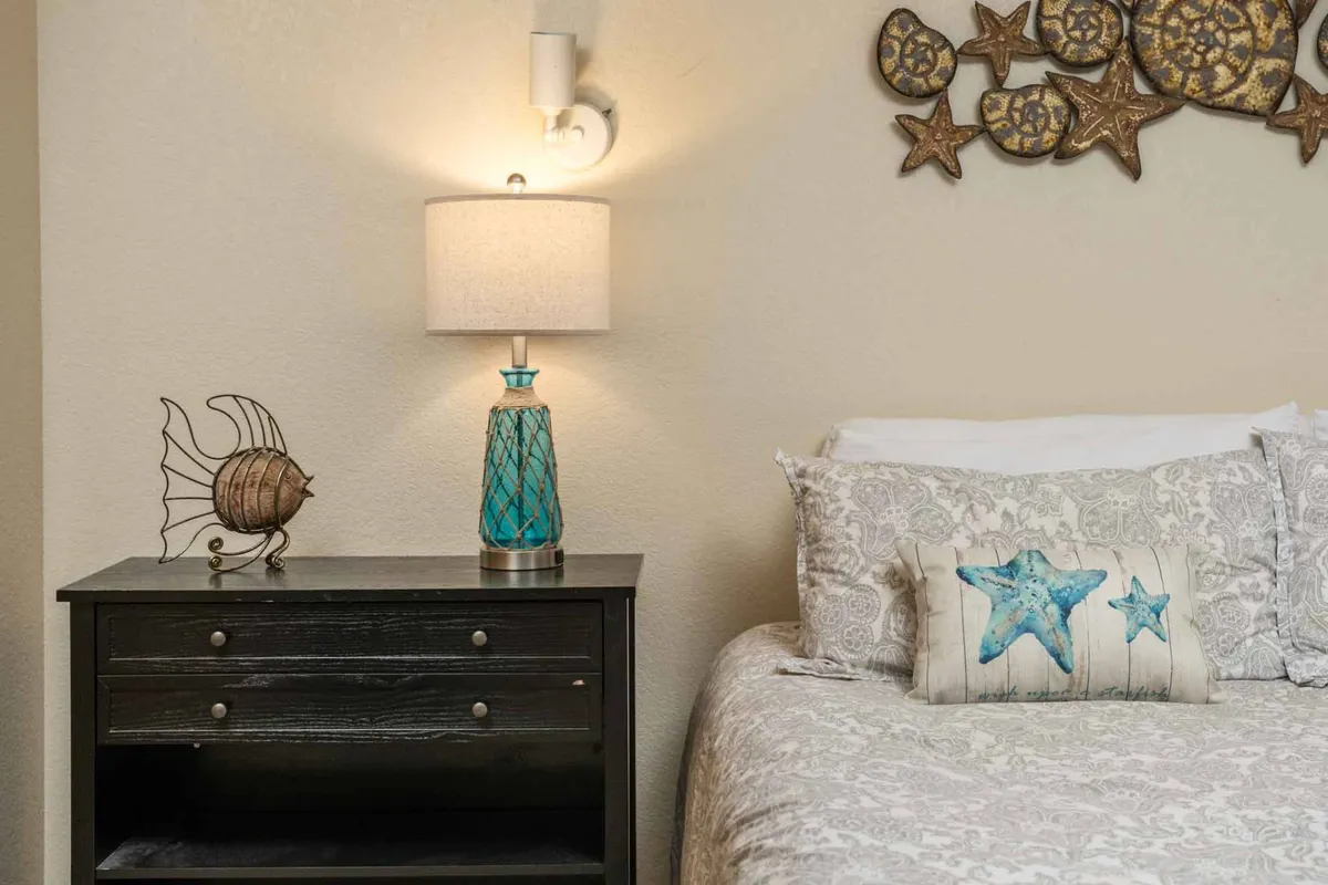 A tranquil bedroom setting with a cozy bed, decorative pillows, a nightstand with a lamp, and a whimsical fish sculpture accent. The space exudes a sense of comfort and calm, ideal for relaxation or bedtime reading.