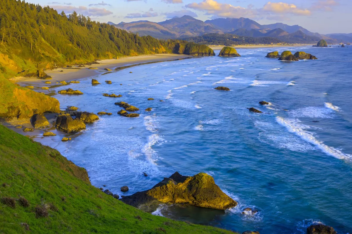 This view captures the dramatic ocean waves rolling onto a wide sandy beach, framed by iconic rock formations and distant mountains. The scene is tranquil, with hints of wildlife and adventure awaiting along the shoreline.