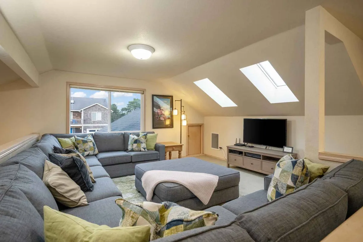 A sunlit living room with a large sectional sofa, skylights, and a television. Decorative pillows and ample natural light create a cheerful, welcoming atmosphere. The layout encourages relaxation, movie nights, and friendly socializing.