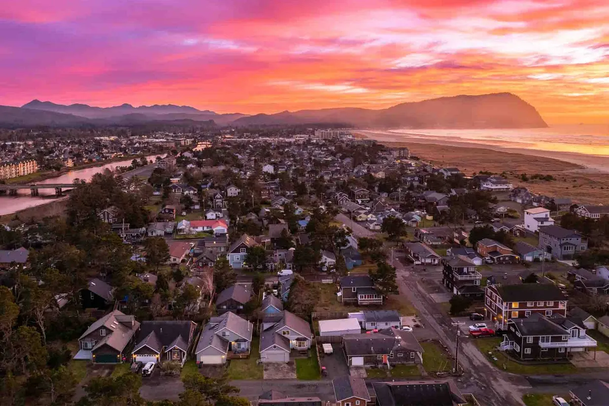 A sweeping view of a vibrant sunset sky over the river winding through the town, with houses nestled among trees and mountains in the distance. The scene captures the inviting character of the Seaside area and its proximity to engaging waterfront attractions.